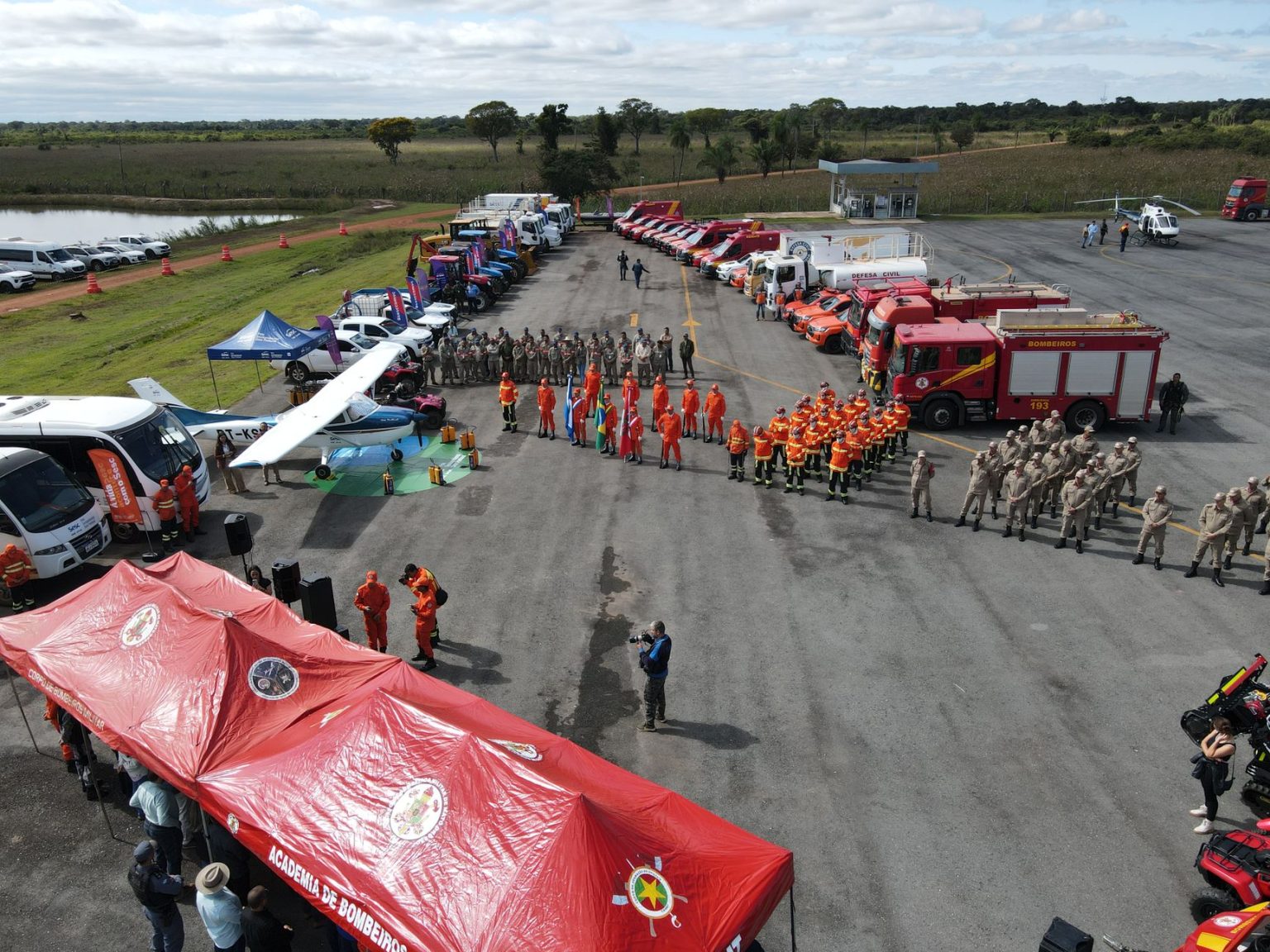 Vista aérea de equipes de combate a incêndios reunidas ao lado de caminhões, equipamentos e tendas em uma área aberta.