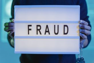 Mãos seguram uma placa com a palavra “fraude”, iluminada por uma luz azul.