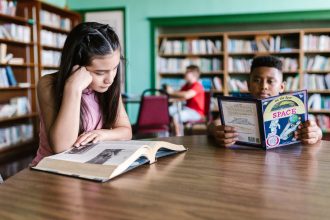Crianças leem juntas à mesa de uma biblioteca, cercadas por livros e estantes.