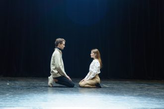 Dois atores participam de uma cena teatral sob um foco de luz no palco.
