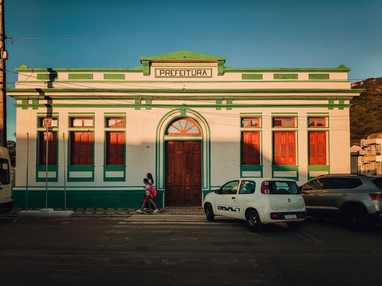 Fachada colorida de uma prefeitura brasileira, com pessoas circulando em frente ao prédio ao entardecer