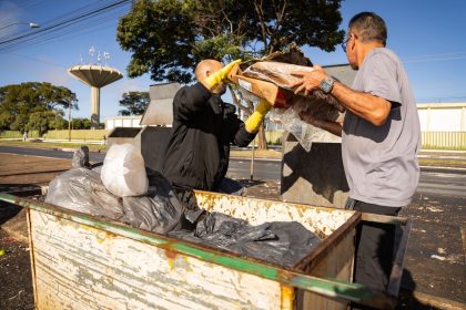 Trabalhadores da limpeza urbana esvaziam lixeiras em uma área urbana sob céu claro.
