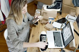 Mulher fotografa sapatos para venda on-line em um espaço de trabalho doméstico, com computador e embalagens ao lado.
